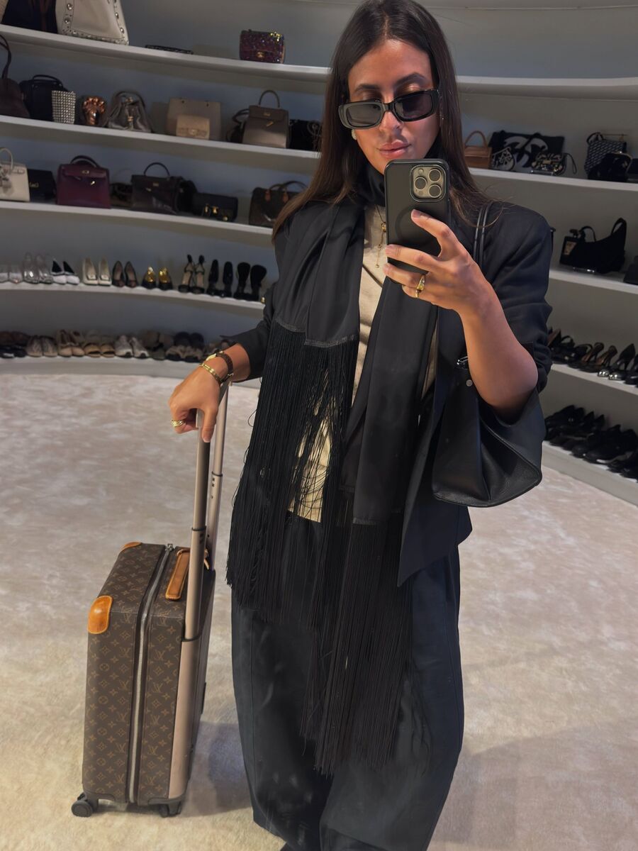 A woman in a black fringed wrap and sunglasses, beside a suitcase