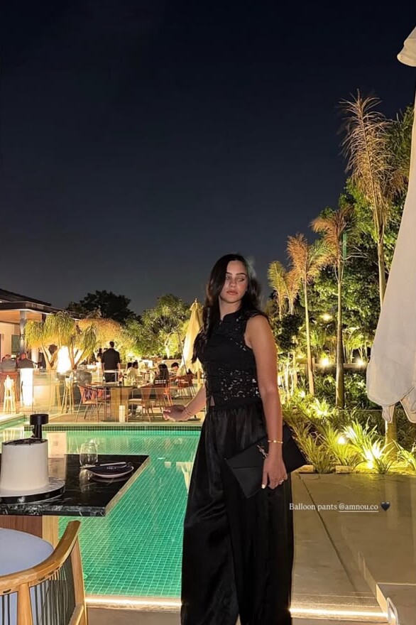 A woman in black lace and black balloon trousers by a lit pool at night