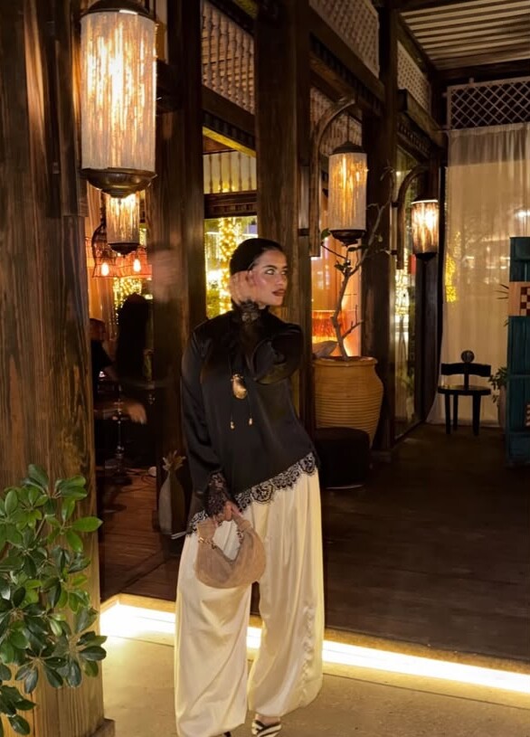 A woman in a black satin blouse with lace and cream balloon trousers, under lanterns at night