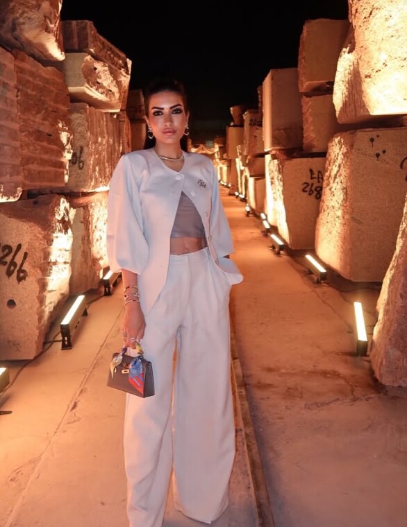 A woman in a white jacket and wide trousers between lit stone blocks at night