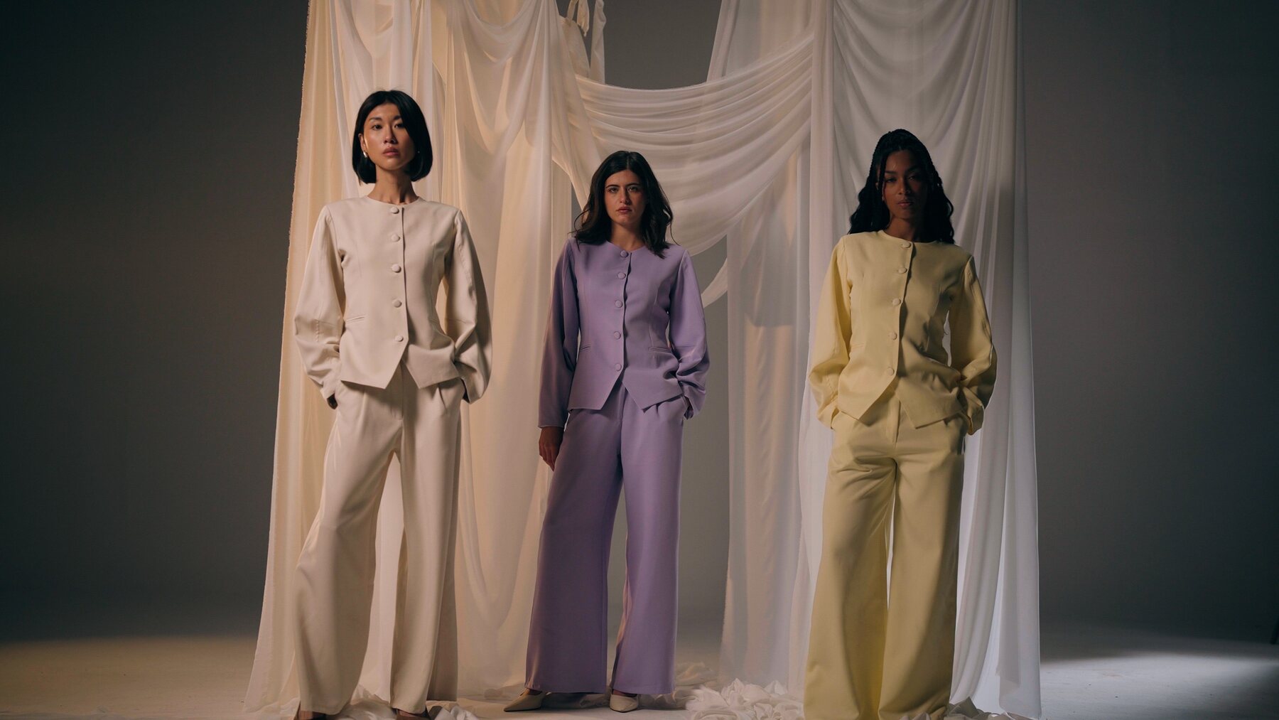 Three women in amnou sets, creamy, lilac and butter yellow, standing before draped fabric
